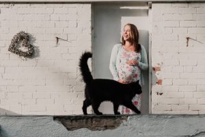 a woman holding her pregnant belly and laughing as a cat photobombs the image