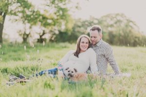 a man and woman sat on the grass in a field with the sun setting behind them and a dog on the floor in front