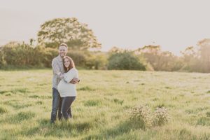 couple stand in field hugging and touching a pregnancy woman's stomach with the spring setting sunlight behind them