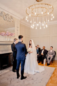 a bride and groom marry in the Pimlico Room of London's Marylebone Town Hall. Photo is taken so you can see the chandelier and white decor of the room