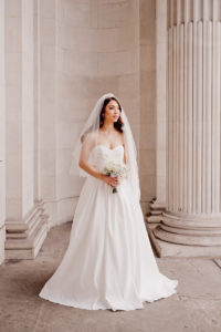 Bride wearing a white wedding dress, white veil and holding a white flower bouquet amongst the arches of London's Marylebone Town Hall