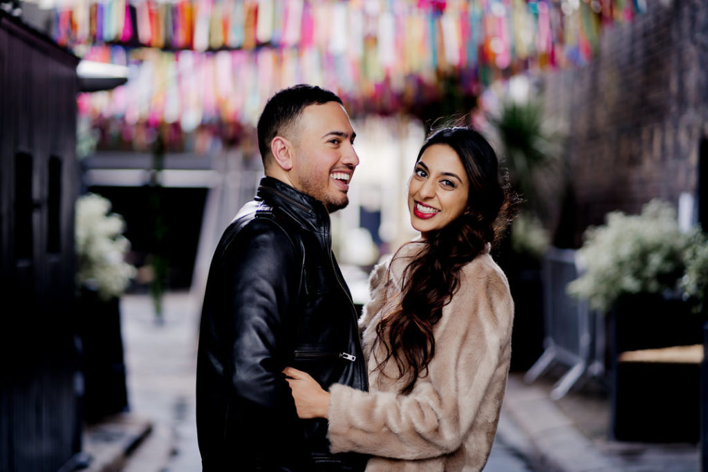 man and woman stand chest to chest both laughing with colourful ribbons hanging in the background of their engagement photo
