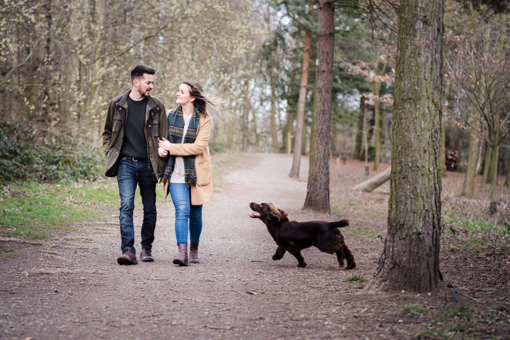 couple walk arm in arm and look at each other smiling while their dog runs around them on an engagement photography session in a wood