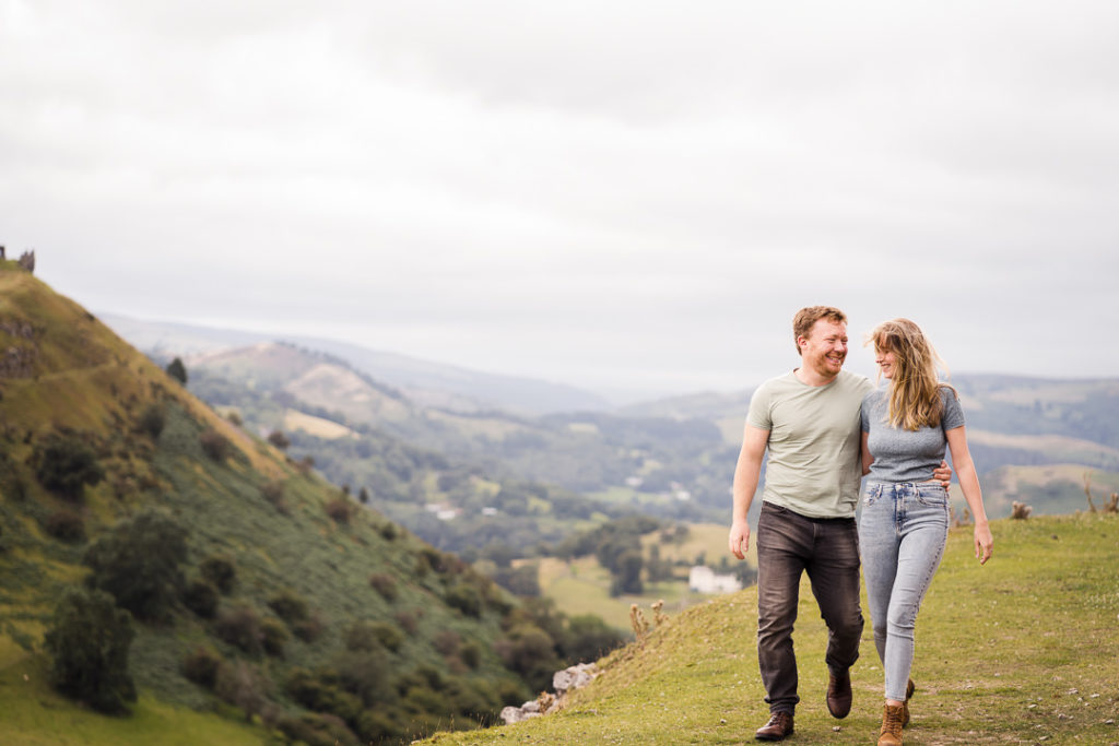 a man and woman walk with their arms around each other and look at one another and smile while on a hill top with gorgeous views behind them in North Wales on their engagement photo session