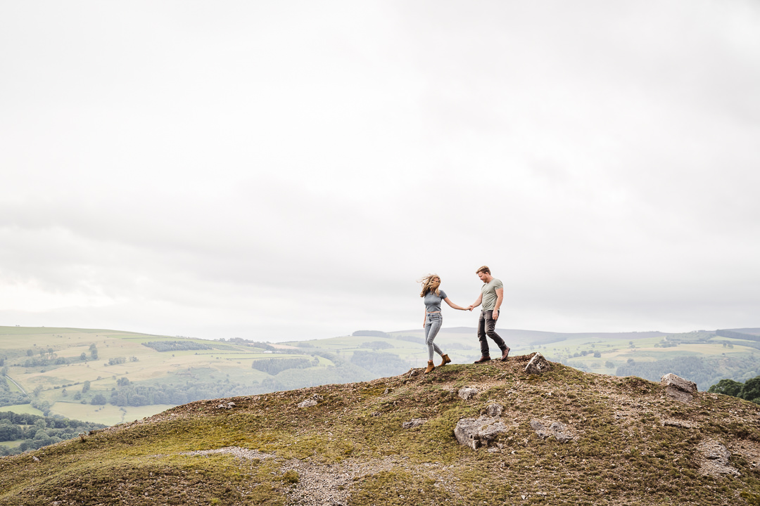 a woman leads a man down a hill while walking and the wind blows her hair so it covers her face and she looks back at him in North Wales on their pre-wedding photography session