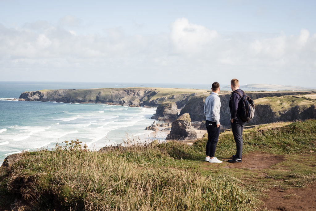 two men stand looking out at an amazing view across a Cornish beach for their engagement photography session