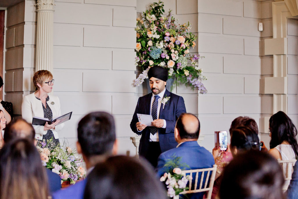 a best man reads a speech as part of a wedding ceremony while the second photographer captures it from the back of the room