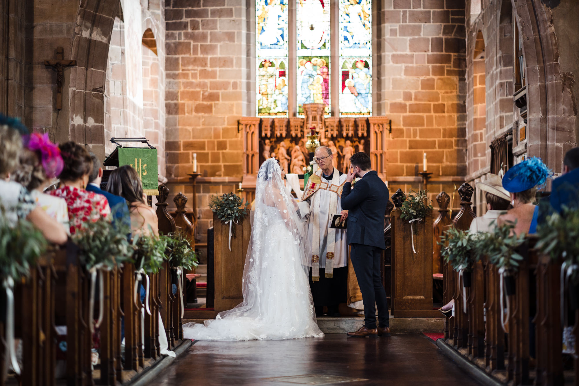 full length of bride in a white dress and veil and groom in blue suit in a church at the alter as they marry
