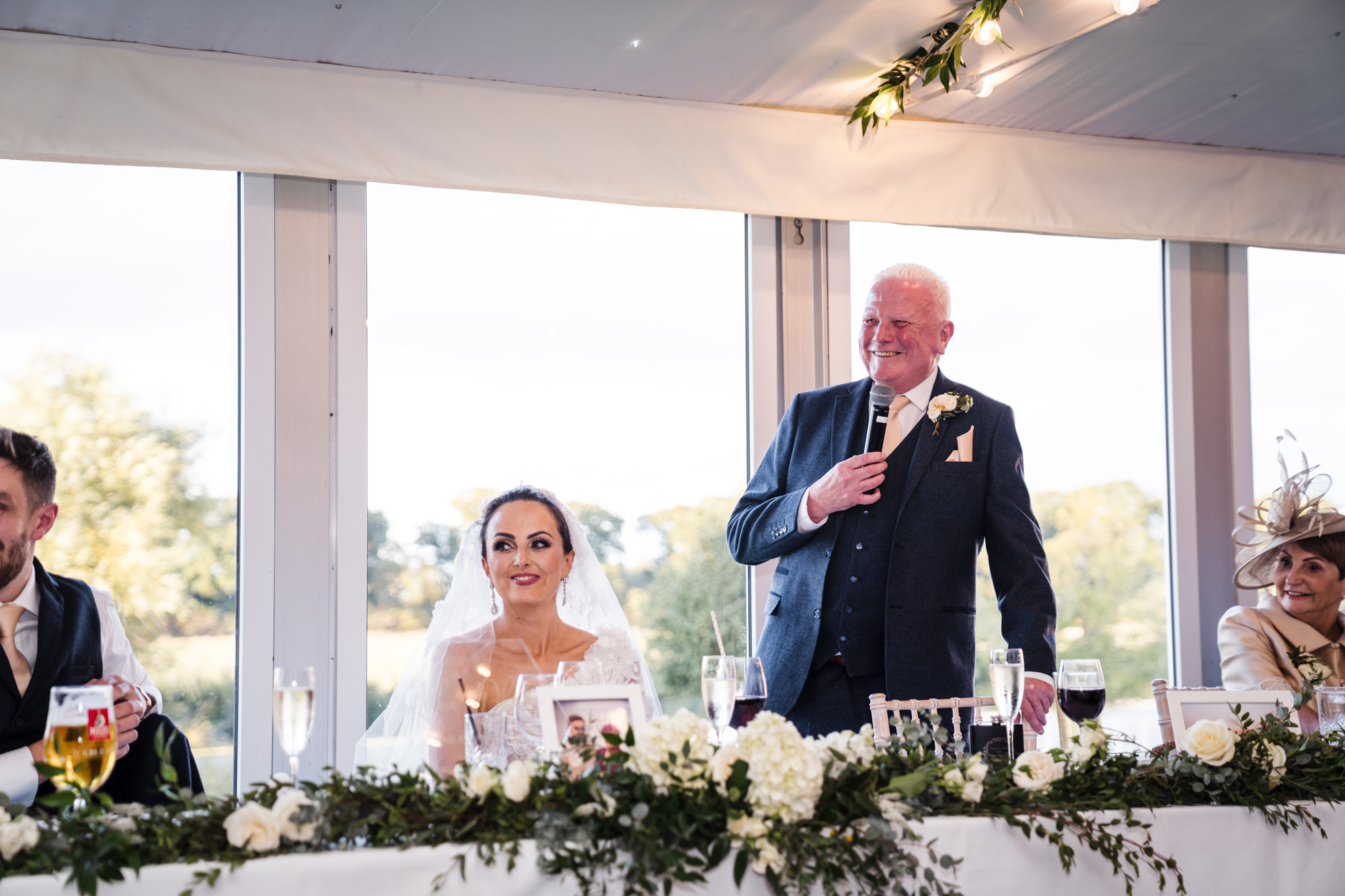 father of the bride giving a speech at wedding while bride smiles