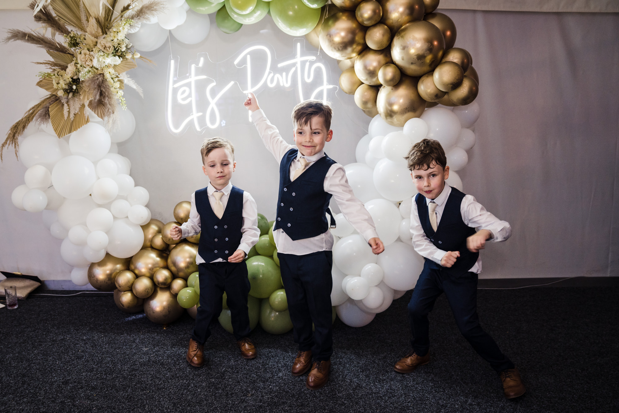 three children act silly at wedding in front of balloon arch and neon lets party sign