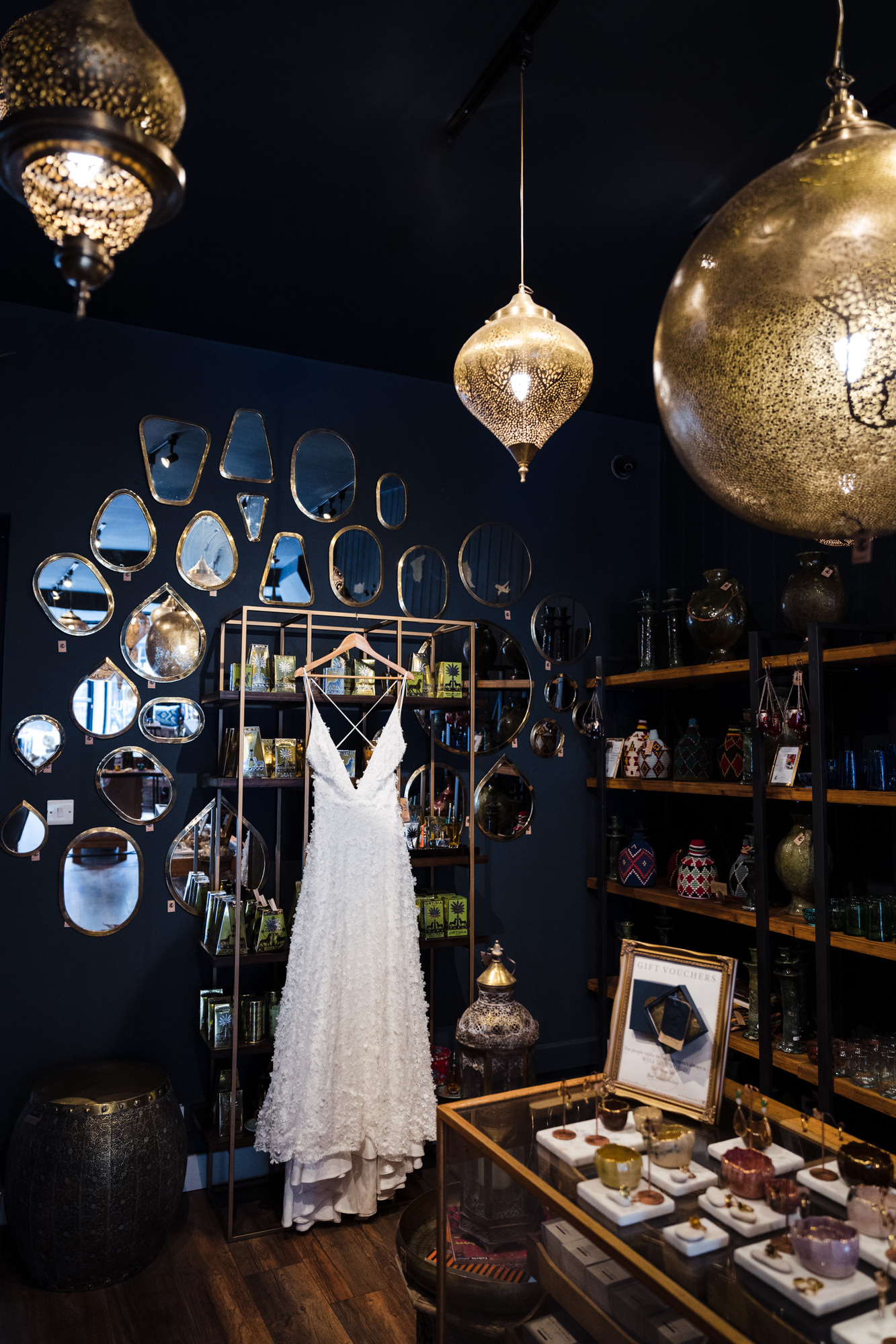 a Charlie Brear luxury wedding dress hangs in a room full of mirrors and gold lamp shades