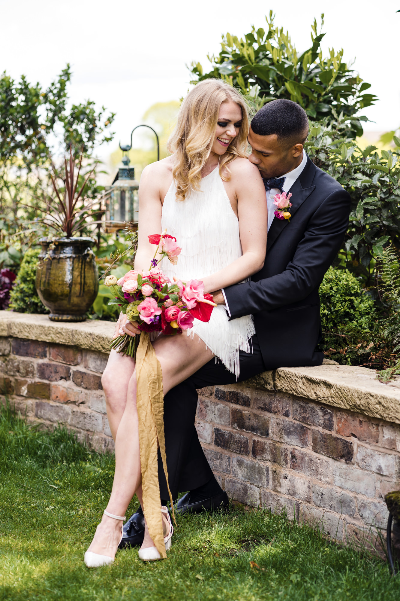 groom sits on a wall while bride sits on his lap and he tenderly kisses her shoulder. bride is holding a bold colourful floral bouquet