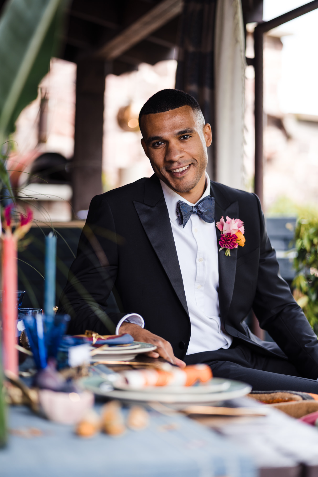 solo portrait of groom sat at table smiling and looking at the camera wearing a black tie tux