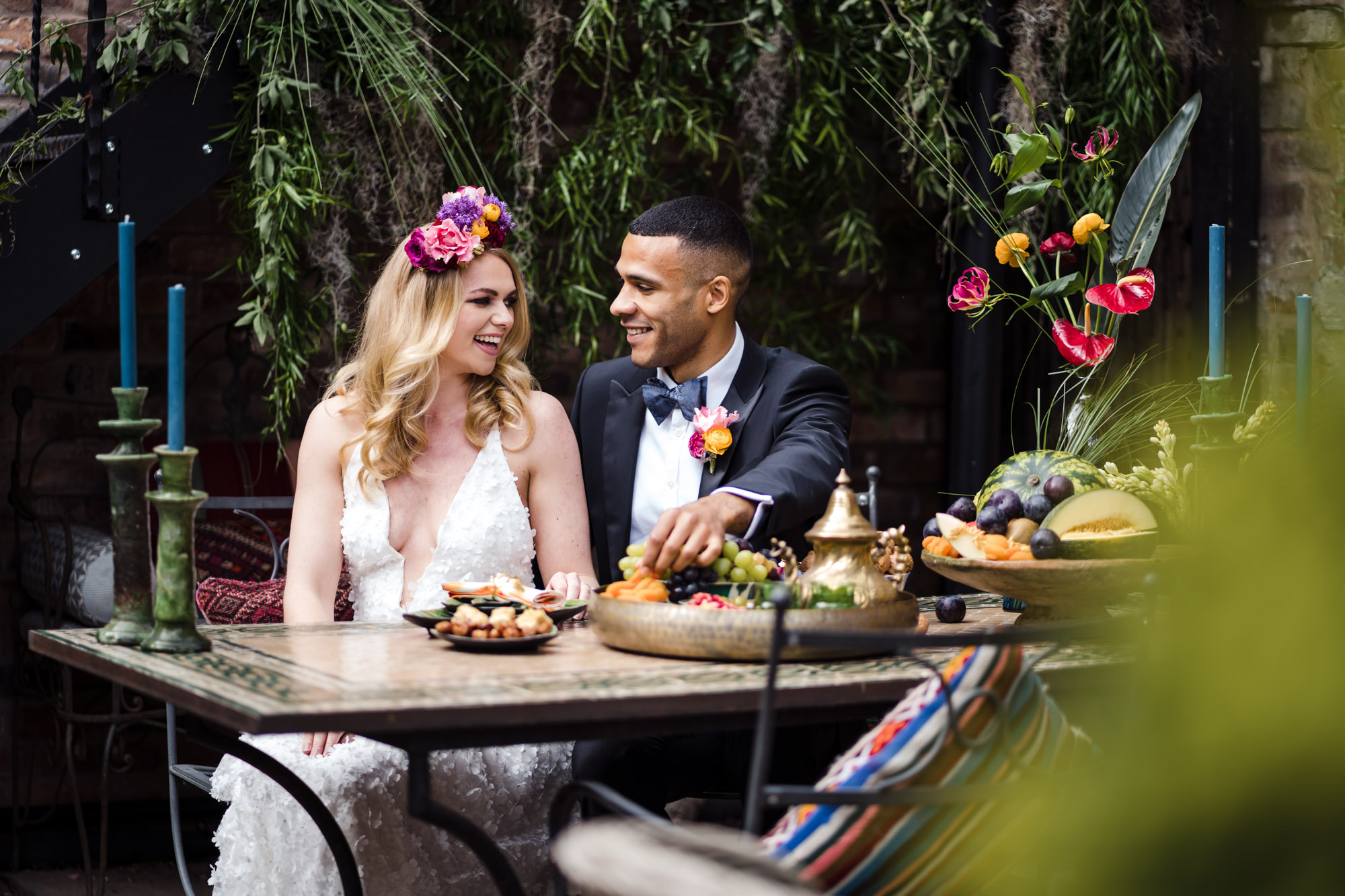 bride and groom sit and feed one another in front of a fruit medley