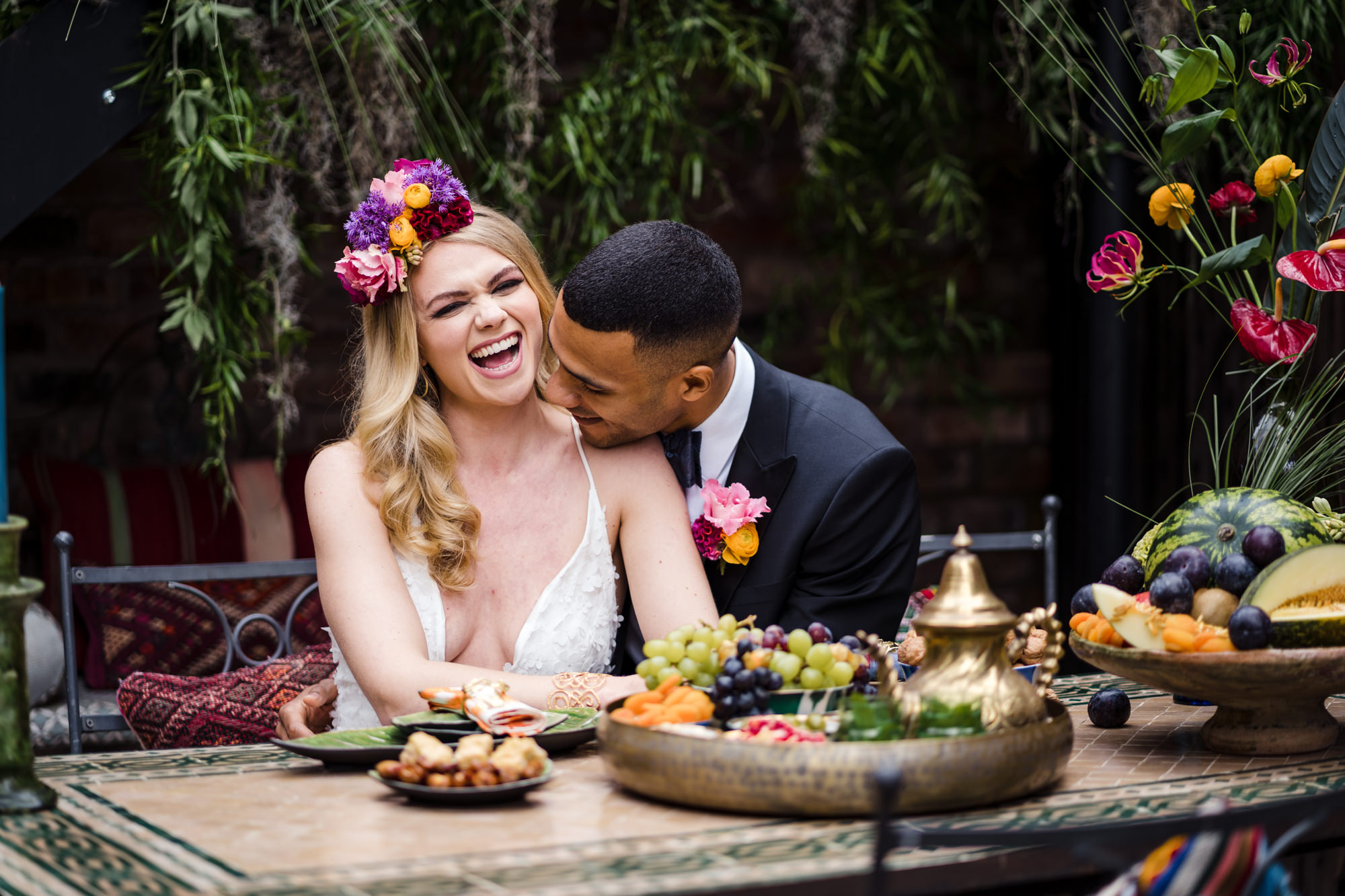 groom kisses bride on the shoulder making her laugh. bride wears a bold colourful statement flower crown