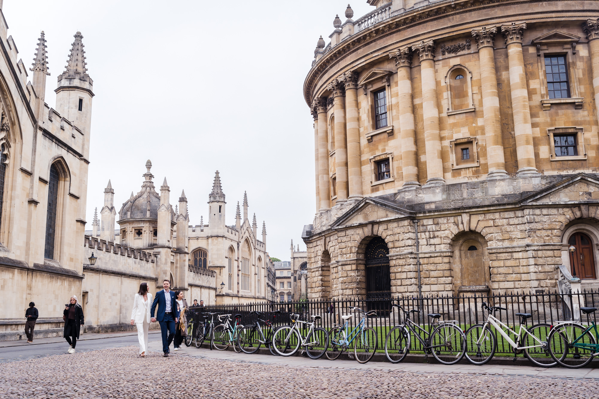 wide shot of bride and groom walking hand in hand looking relaxed past Radcliffe camera