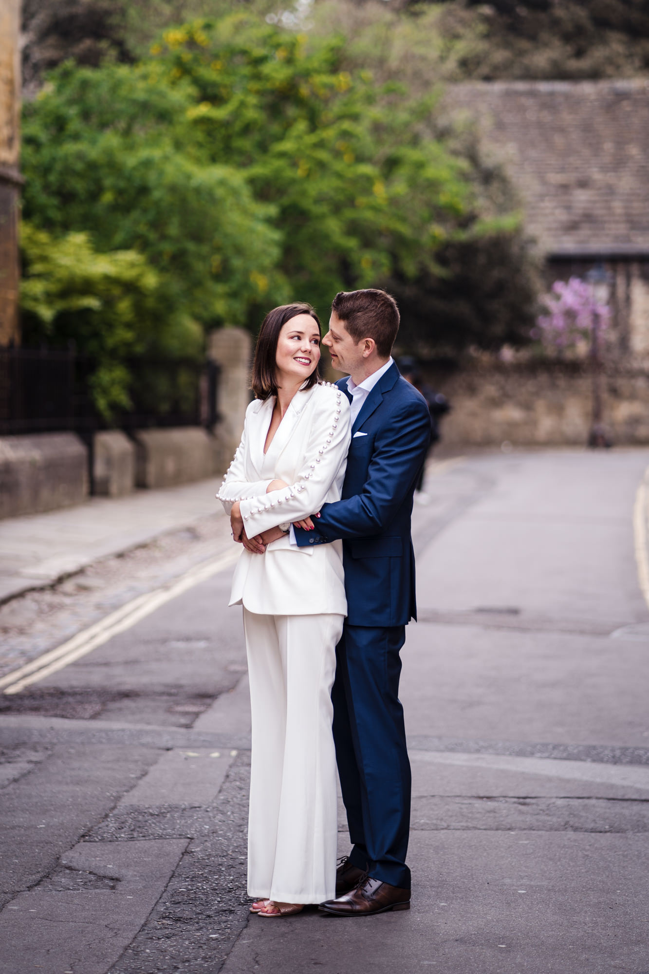 bride wears a white suit and hugs groom in blue suit for couple portrait at their oxford registry office wedding elopement