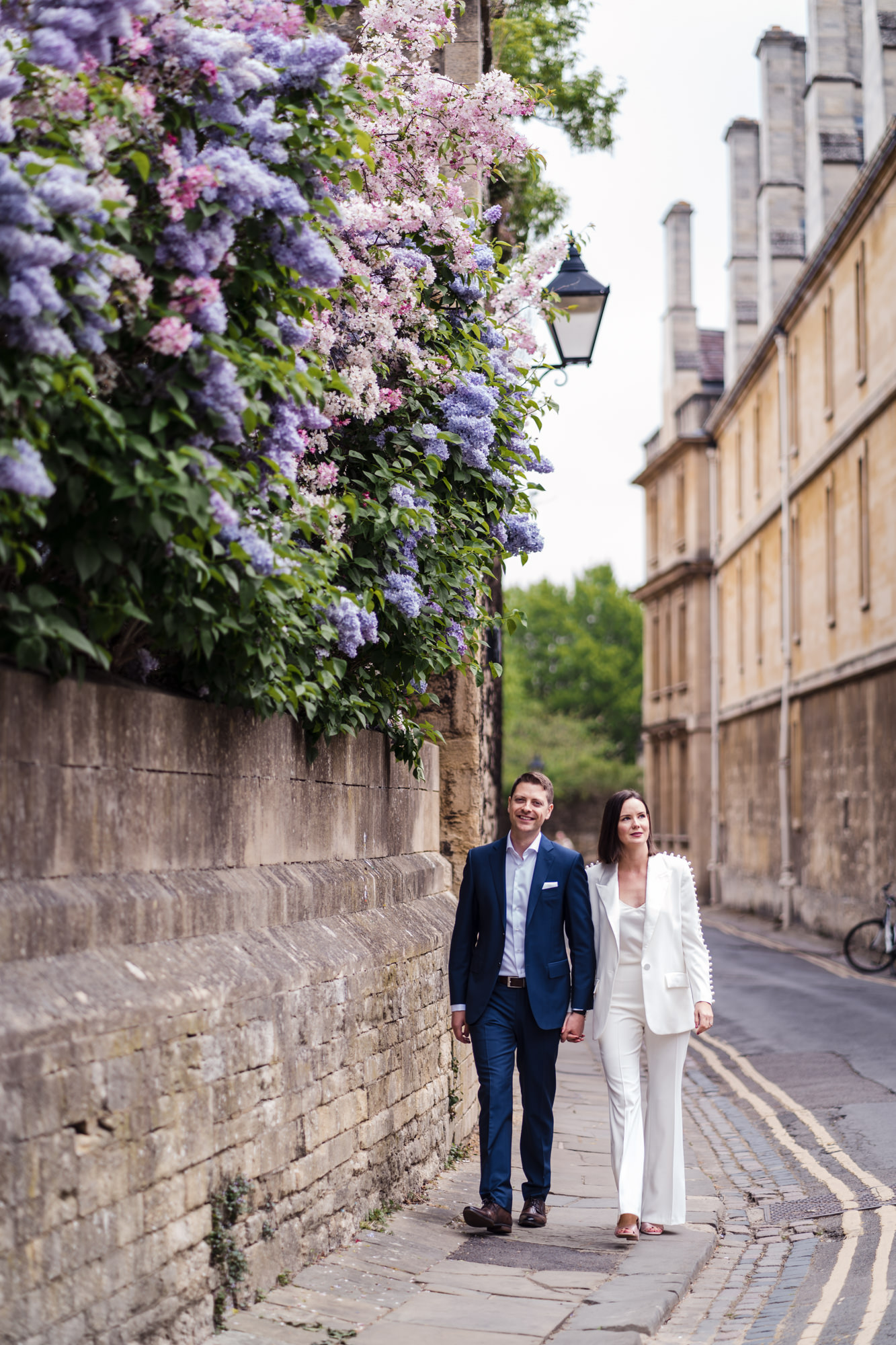 bride and groom walk alongside a wall with purple and blue flowers for modern couple wedding portrait in oxford city