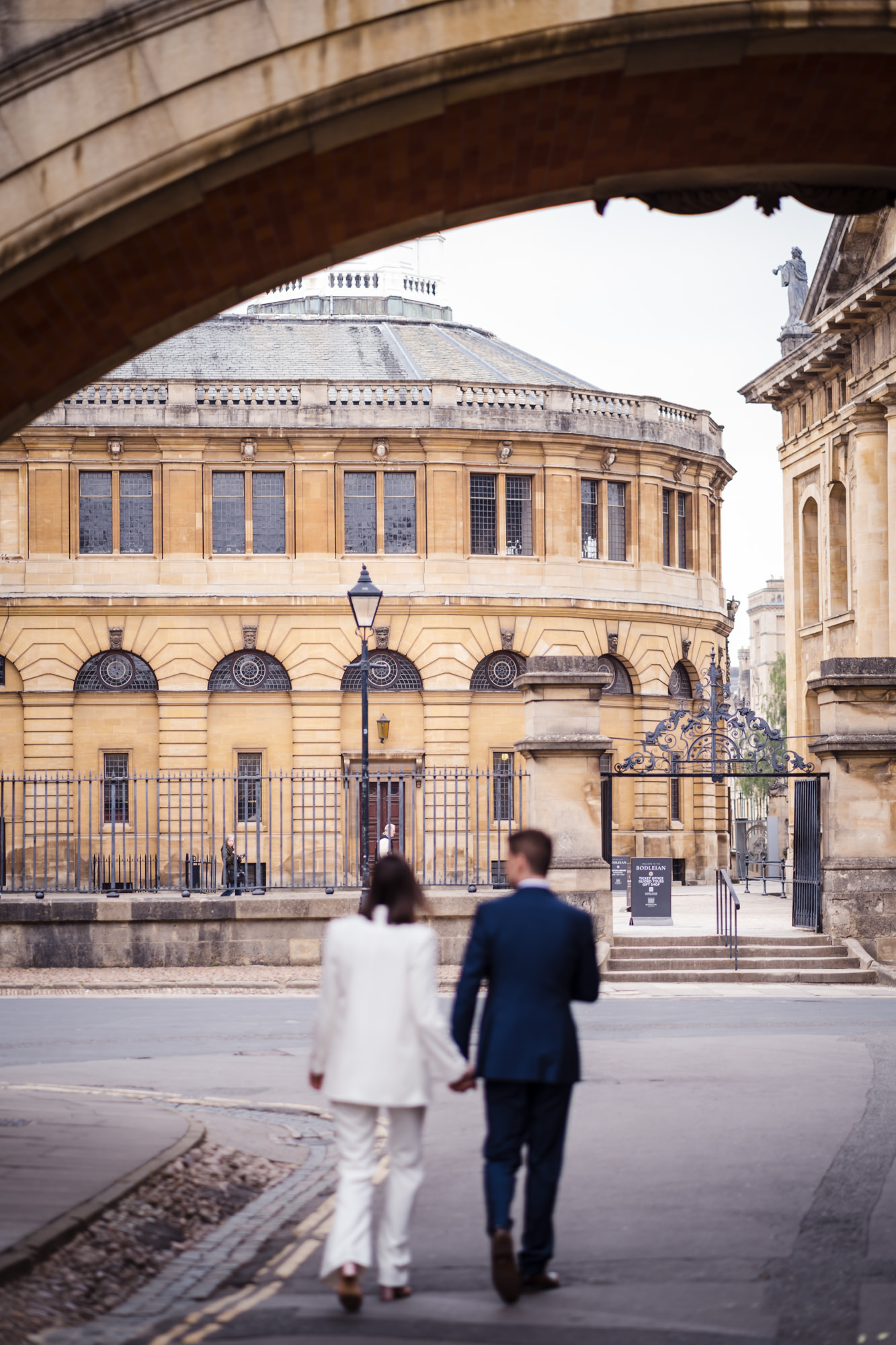 bride and groom walk hand in hand underneath the bridge of sighs with Bodleian Library oxford in focus