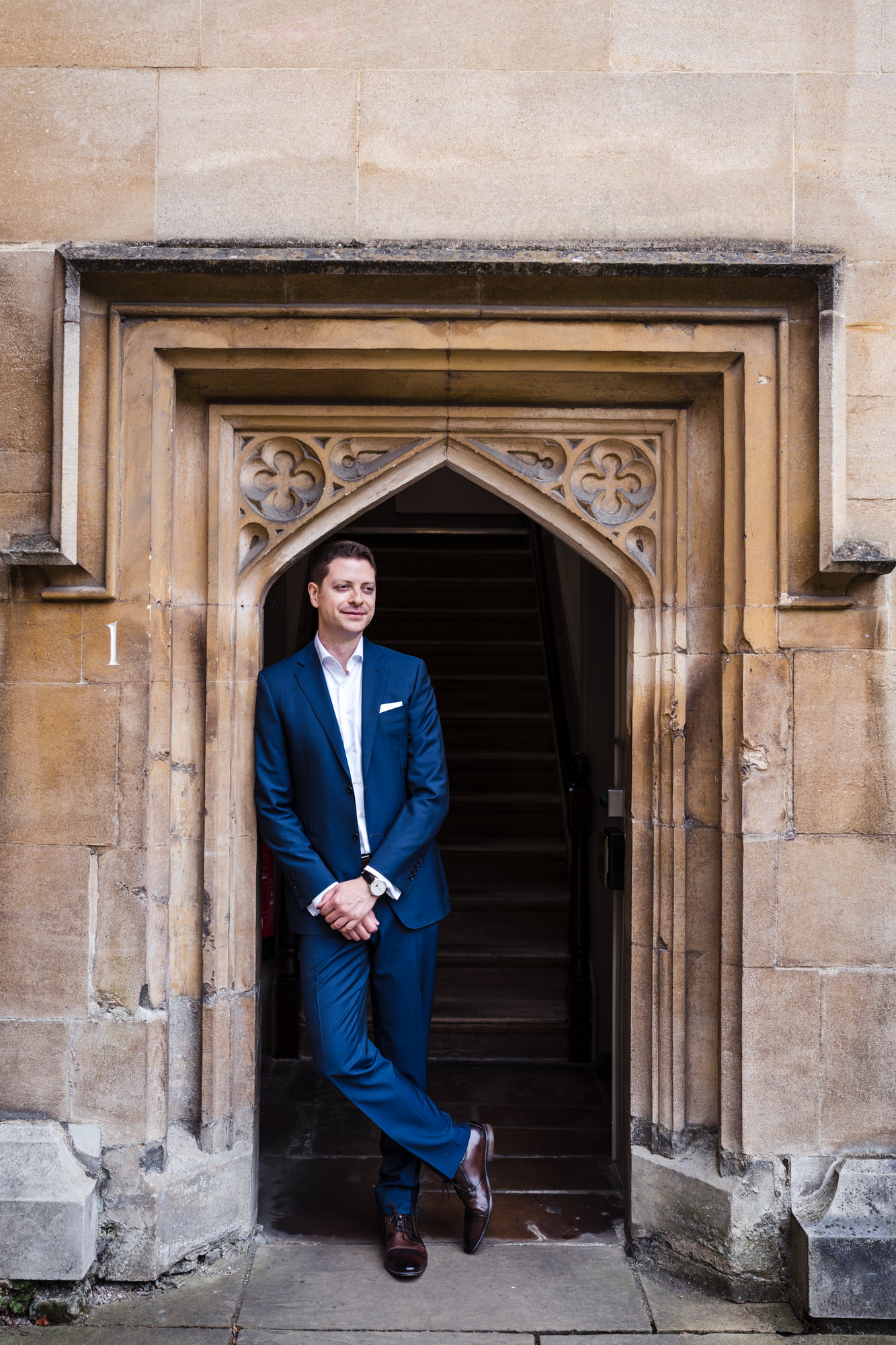groom in blue suit leans against a doorway at lincoln college oxford at his small wedding elopement