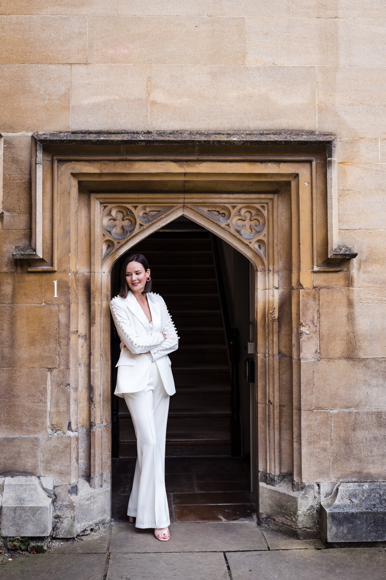 bride leans against wall at Lincoln College Oxford wearing a white Nadine Merabi Bridal Suit