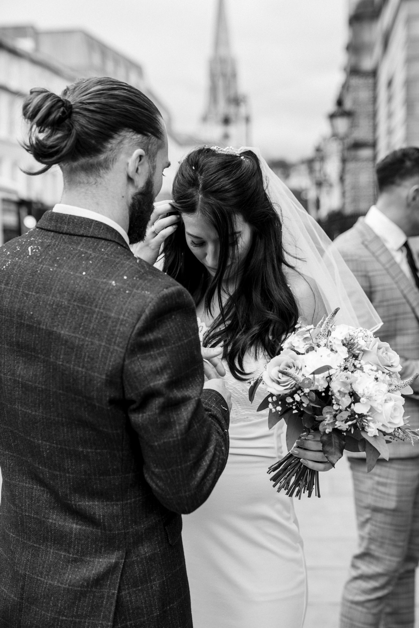groom helps bride brush off confetti from her dress in black and white image as bride looks down while holding a bouquet