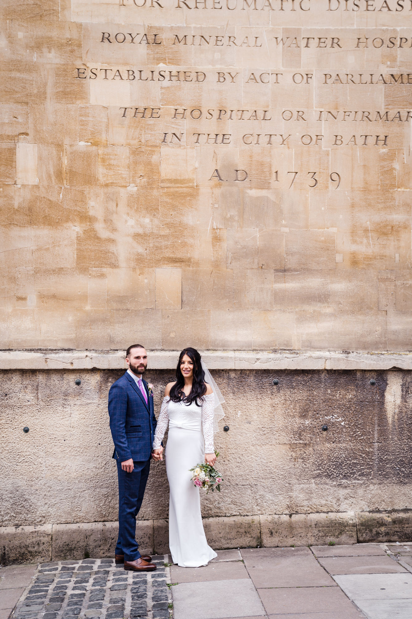 couple stand side by side and look at the camera while stood in front of a bath stone wall