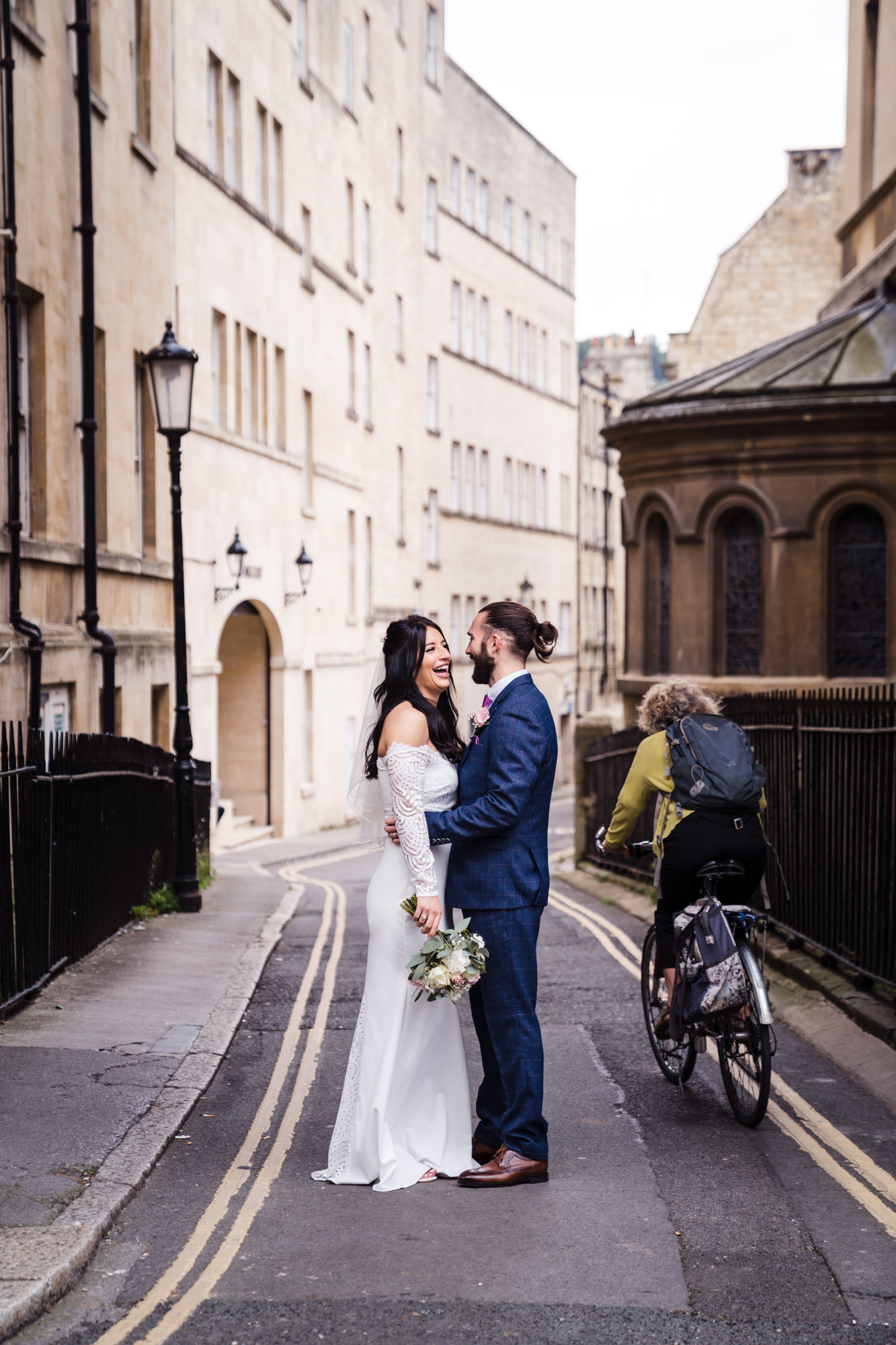 cyclist passes bride and groom as they pose for couple portrait in Bath and bride laughs at the situation