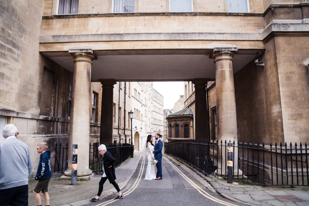 wide city shot of bride and groom as passers by cross in front of the photo in bath