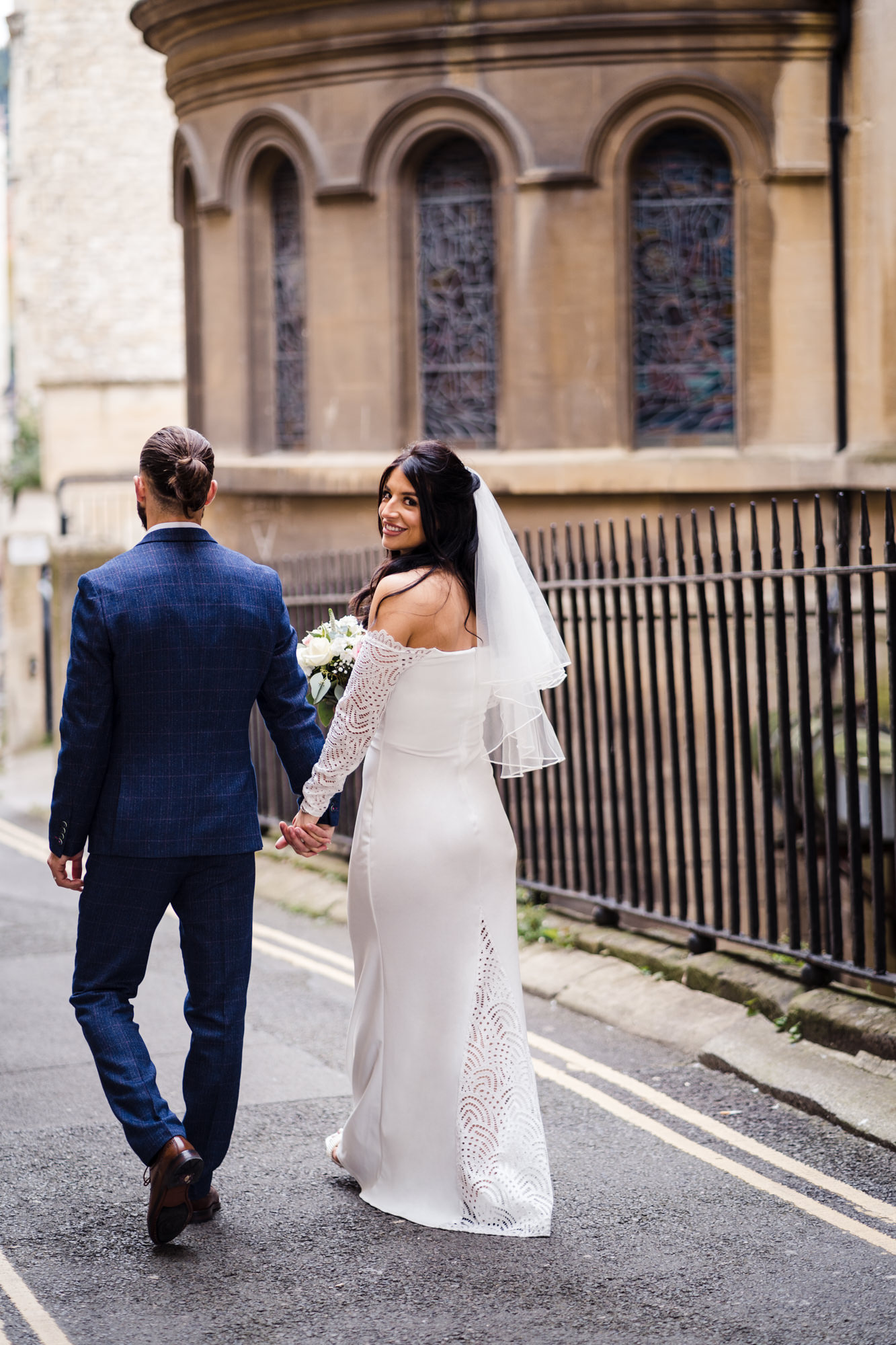 groom and bride walk through Bath city as bride looks over her shoulder and smiles at the camera