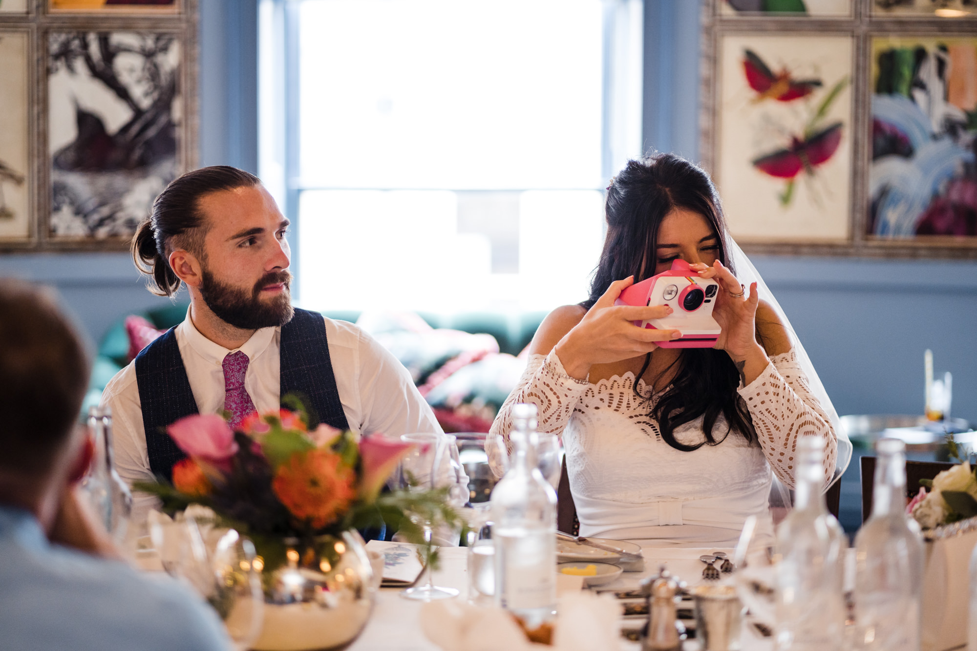 groom and bride sit at the table waiting for wedding breakfast while bride snaps a photo with her Polaroid camera