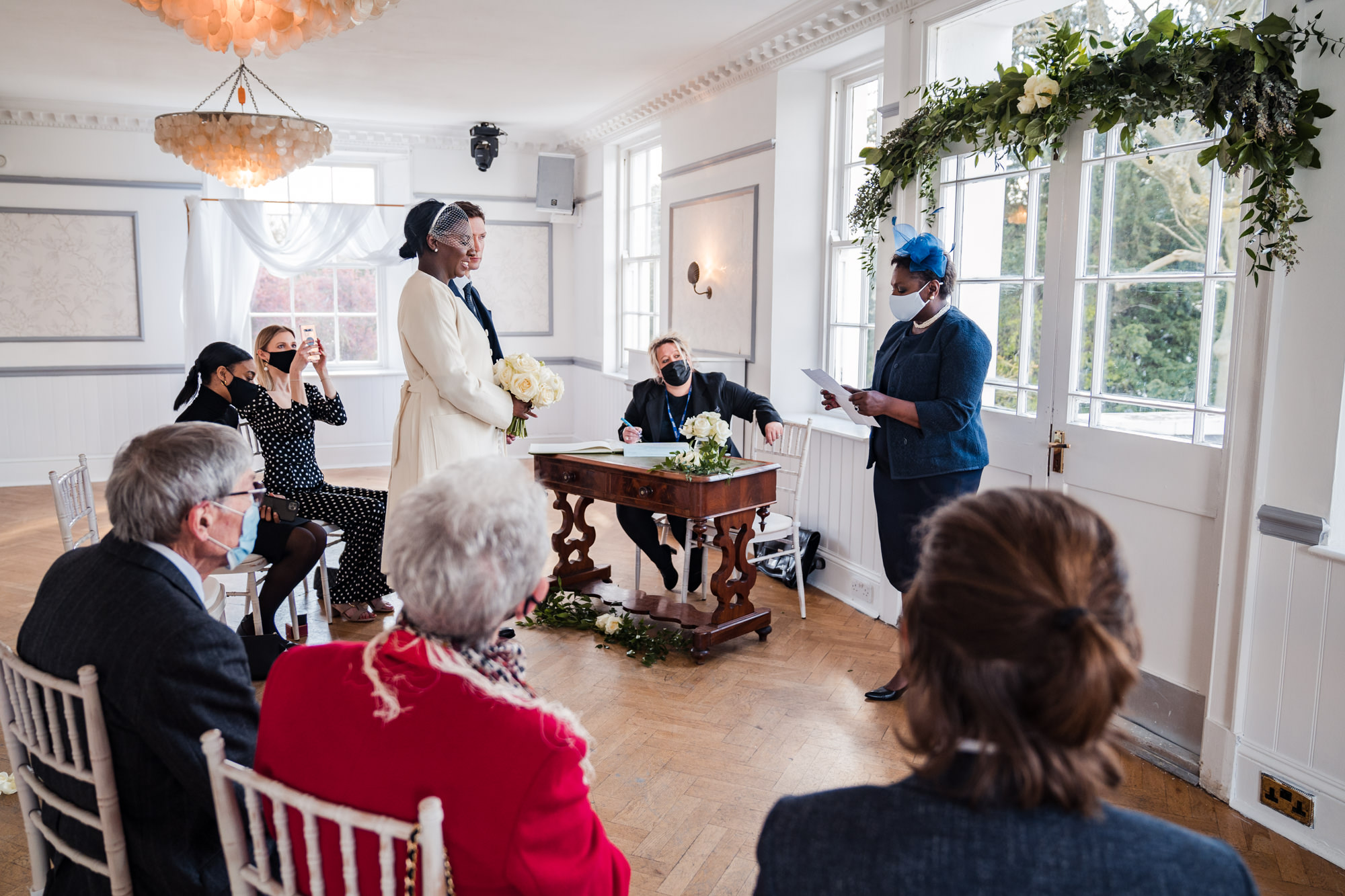 wide shot of wedding ceremony as mum reads a reading, bride and groom look on and a guest films on her phone
