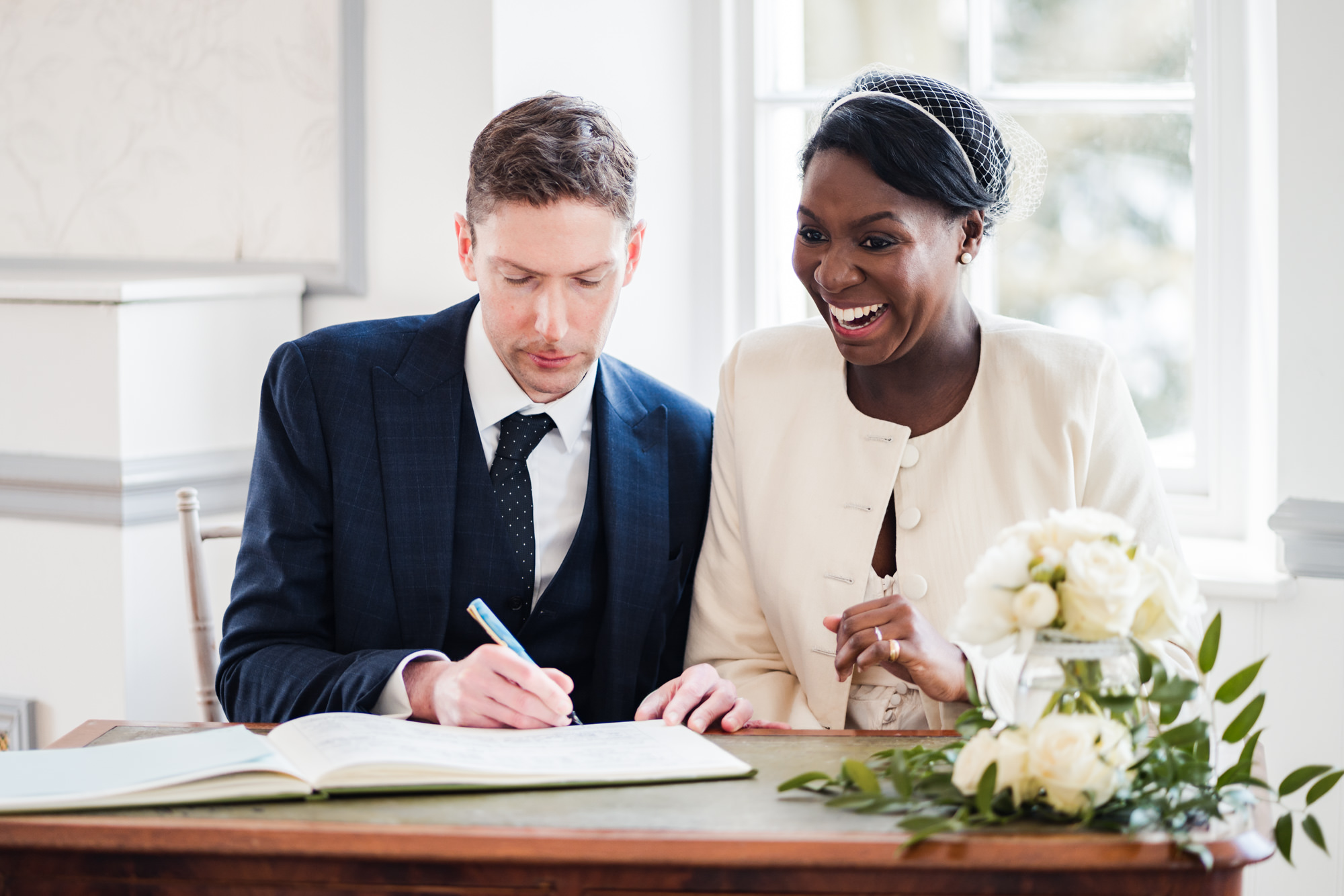 happy bride smiles in delight as groom signs wedding register during their wedding ceremony