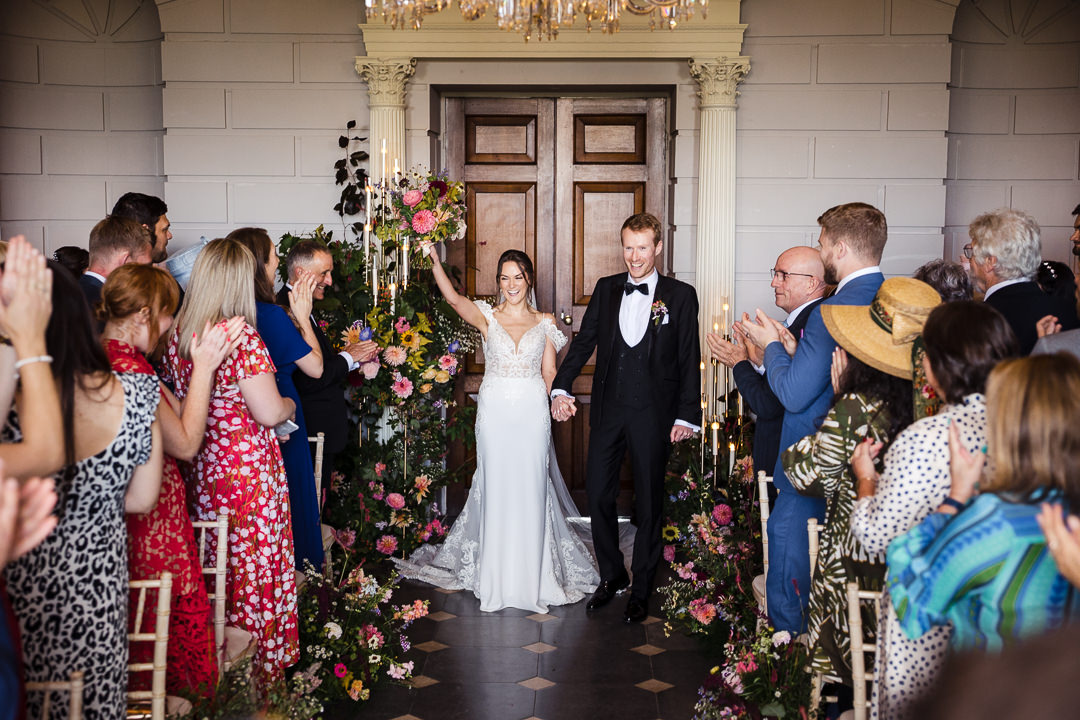 Just married bride raises her hand in celebration as the room applauds, framed by colourful floor aisle meadow flowers and candles