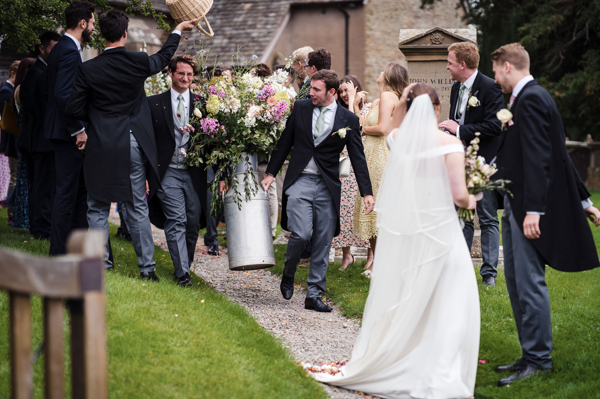 Groomsmen carry tall English-garden floral urns from the church as the bride watches, just after the church ceremony in Shropshire amid guests laughing