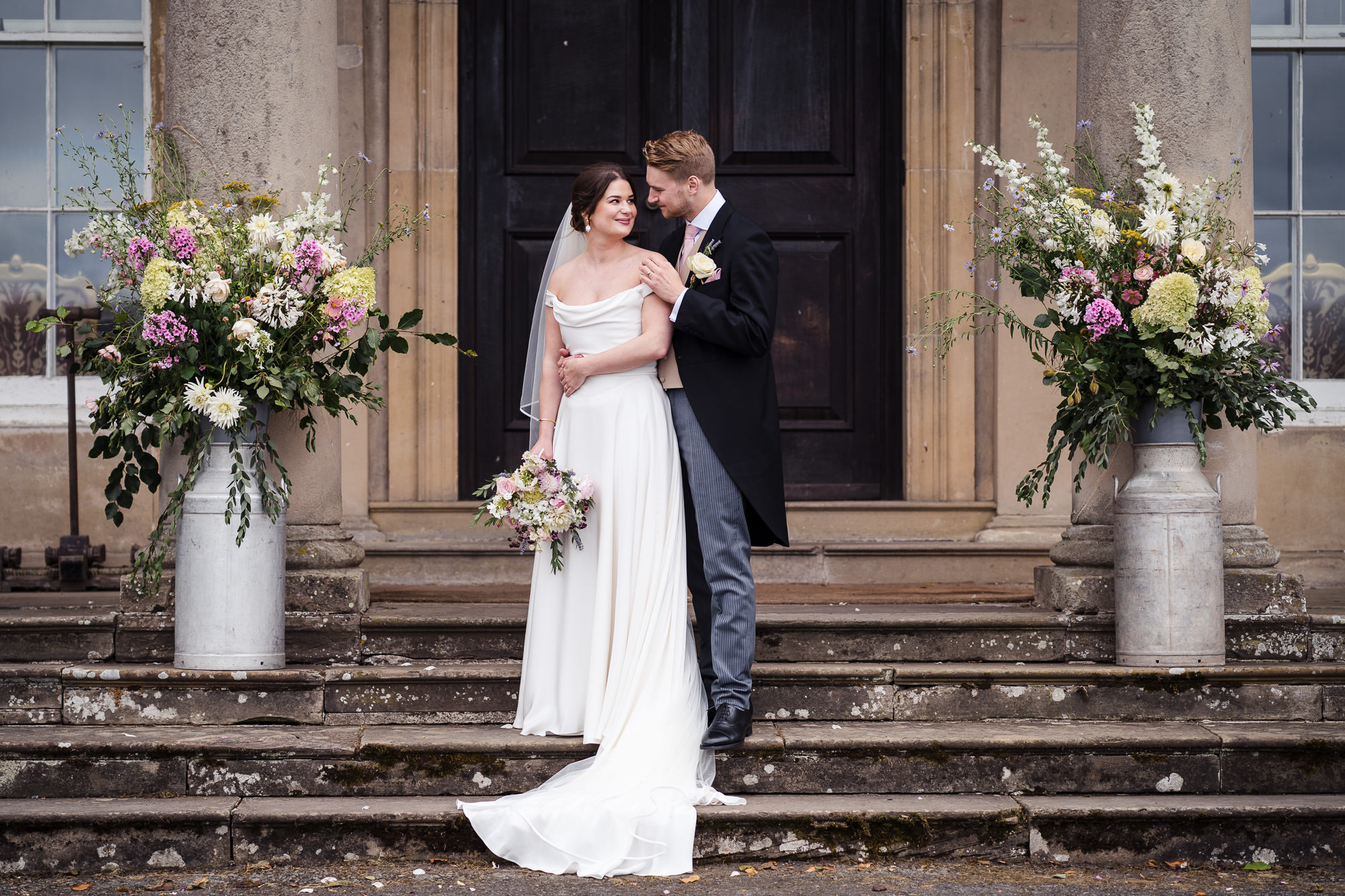 Couple on Walcot Hall steps between tall urns of pastel garden flowers; Halfpenny London dress and veil