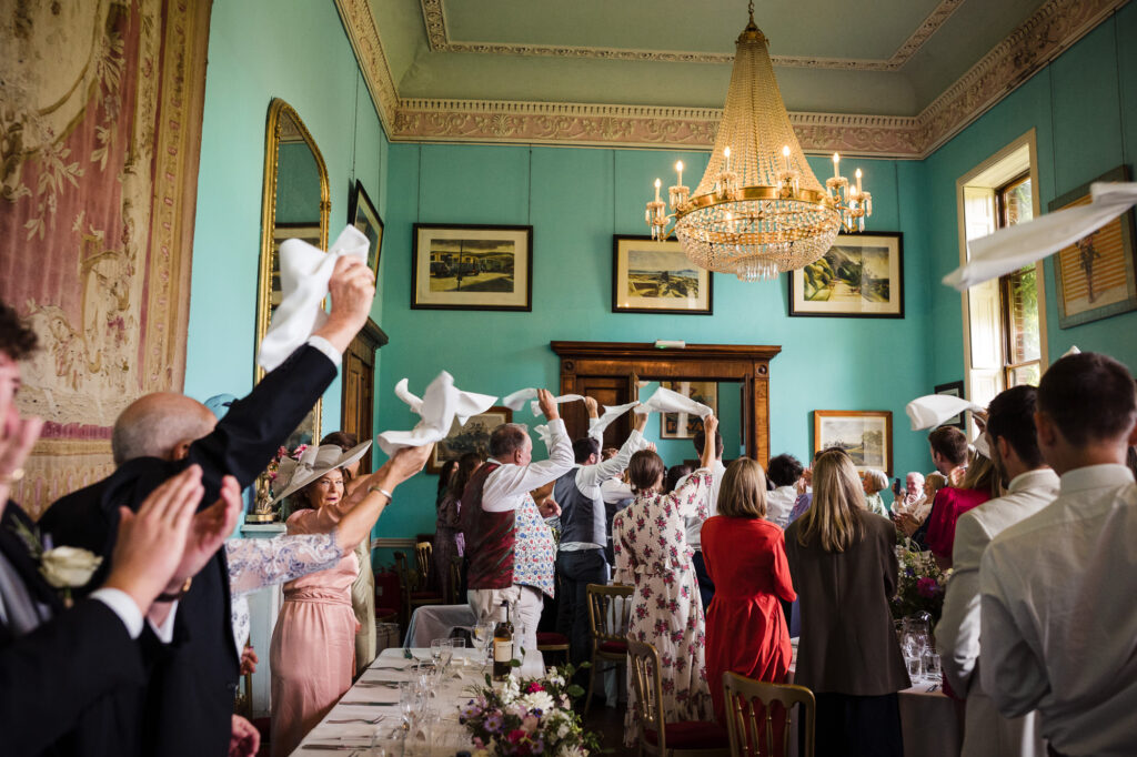 Wedding breakfast entrance at Walcot Hall - guests cheers and wave napkins under the crystal chandeliers in the teal dining room