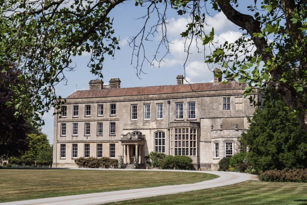 Elmore Court manor house exterior with sweeping lawn and curved driveway, framed by leafy trees.