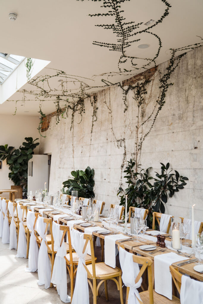 Long banquet table setup at Middleton Lodge with wooden chairs, greenery overhead, skylights and a concrete feature wall.